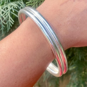 May include: Three silver bangles stacked on a person's wrist. The bangles are smooth and shiny.