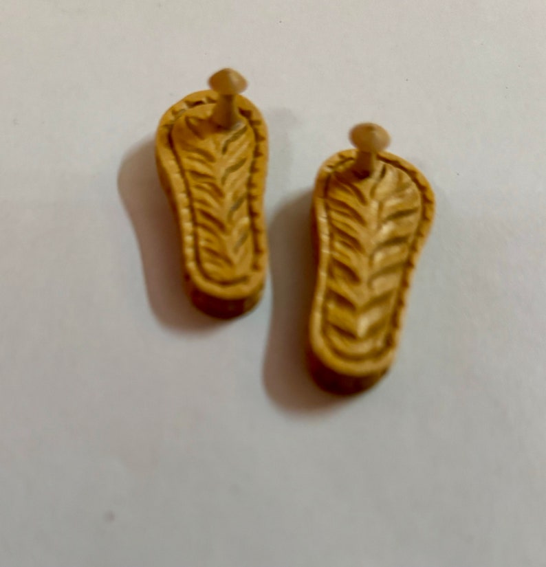 May include: Two wooden sandals with a carved design. The sandals are light brown and have a small wooden peg on the back.