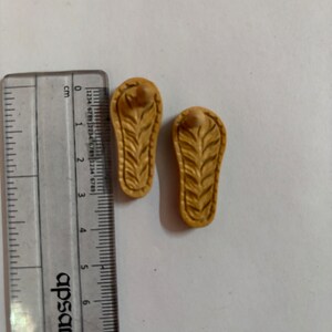 May include: Two small, brown, wooden sandals with a carved leaf design. The sandals are about 2.5 cm long.