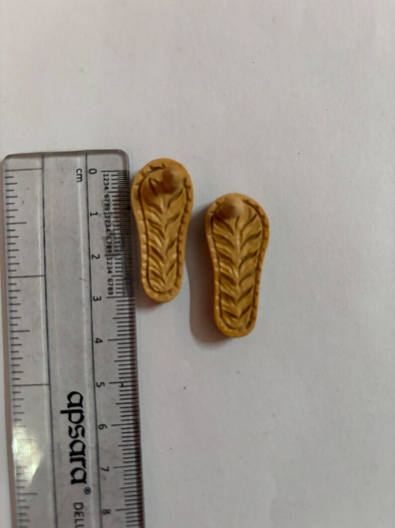 May include: Two small, brown, wooden sandals with a carved leaf design. The sandals are about 2.5 cm long.