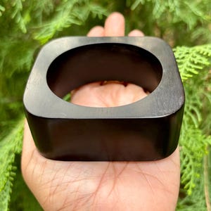 Könnte beinhalten: Ein quadratisches, dunkelbraunes Holzarmband mit abgerundeten Ecken. Das Armband wird in einer Hand gehalten, mit einem grünen, blättrigen Hintergrund. Das Armband hat eine glatte, polierte Oberfläche und eine große Öffnung in der Mitte.