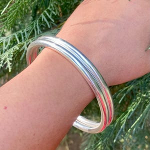 May include: A silver bangle bracelet with a wide, rounded design. The bracelet is made of two thick bands of silver that are joined together.