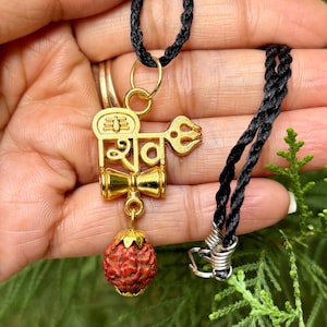 Könnte beinhalten: Eine goldfarbene Anhängerkette mit einem religiösen Symbol und einer roten, strukturierten Perle. Der Anhänger hängt an einer schwarzen Kordel. Die Kette wird in der Hand einer Person vor grünem Hintergrund gehalten.