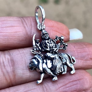 May include: Silver pendant featuring a detailed depiction of a Hindu goddess riding a lion. The goddess is adorned with intricate jewelry and holds a weapon in one hand.