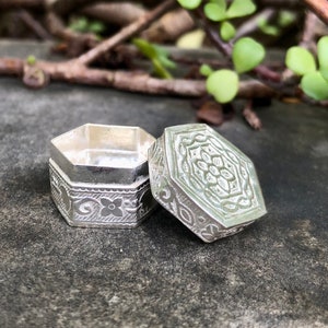 Puede incluir: Una pequeña caja hexagonal de plata con intrincados diseños florales. La caja está abierta, revelando un interior vacío.