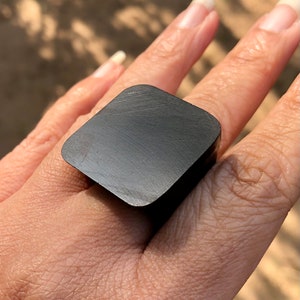 May include: A black square ring with a brushed finish. The ring is worn on a finger.