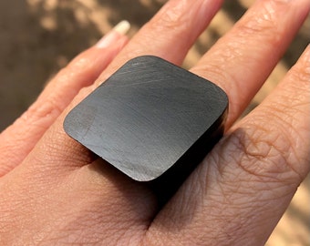 May include: A black square ring with a brushed finish. The ring is worn on a finger.