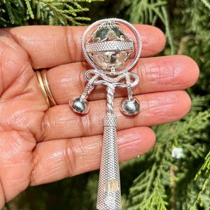 May include: A silver baby rattle with a round, spinning ball at the top and three bells hanging from the bottom. The handle is long and cylindrical with a textured surface.