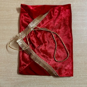 May include: A red velvet pouch with a gold tassel and a gold cord drawstring. The pouch is closed and lying flat on a wooden surface.