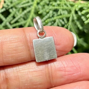 Puede incluir: Un pequeño colgante plateado con una cara rectangular y un lazo para una cadena. El colgante se sostiene entre los dedos sobre un fondo verde borroso. El colgante tiene una superficie texturizada.
