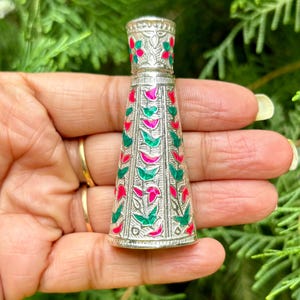 May include: A silver perfume bottle with a conical shape. The bottle is decorated with red, green, and pink floral patterns. The bottle is held in a hand against a backdrop of green foliage.