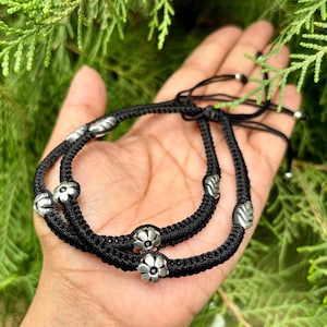 May include: Two black braided anklets with silver flower-shaped charms. The anklets are tied together with a knot.