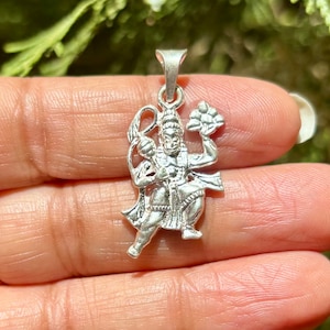 May include: Silver pendant featuring a detailed depiction of the Hindu deity Hanuman, holding a lotus flower in one hand and a club in the other. The pendant is intricately crafted with fine details and a polished finish.