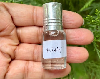 May include: A small, clear glass bottle with a silver cap, held in a hand. The bottle contains a clear liquid and has a white label with the handwritten word "mitti". The background is blurred green foliage.