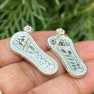 Puede incluir: Un par de dijes de pantuflas de color plateado con intrincados diseños florales. Los dijes tienen un pequeño lazo en la parte superior para sujetarlos a una pulsera o collar.