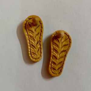 May include: Two small, beige, wooden sandals with a carved leaf design. The sandals have a rounded toe and a small, raised heel.