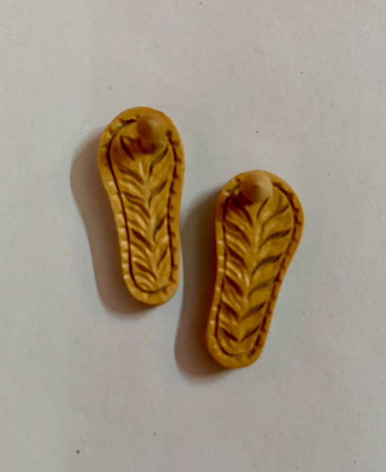 May include: Two small, beige, wooden sandals with a carved leaf design. The sandals have a rounded toe and a small, raised heel.