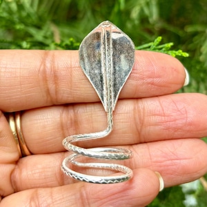 Könnte beinhalten: Ein silberner Ring in Form einer Kobra. Der Ring hat einen Kobrakopf und ein gewendeltes Band. Der Kobrakopf hat ein detailliertes Design, und das Band ist mit einem sich wiederholenden Muster verziert. Der Ring wird in einer Hand vor einem grünen Hintergrund gehalten.