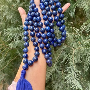 May include: A long necklace of deep blue spherical beads, held in an open hand. A matching blue tassel hangs at the end. The background is a blurred view of green foliage.