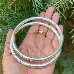 May include: Two silver bangle bracelets, one slightly larger than the other, are stacked on top of each other. The bracelets are smooth and shiny.