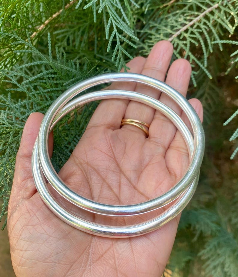 May include: Two silver bangle bracelets, one slightly larger than the other, are stacked on top of each other. The bracelets are smooth and shiny.