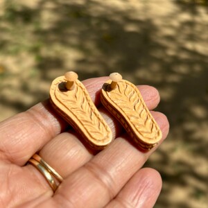 May include: A pair of wooden earrings with a carved leaf design. The earrings are a light brown color and have a post back.
