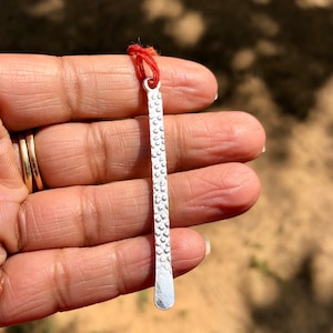 Puede incluir: Un marcapáginas de plata con una superficie texturizada. El marcapáginas está unido a una cuerda roja.