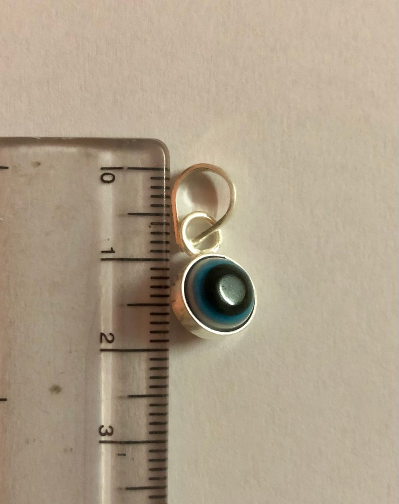 Puede incluir: Un amuleto de plata con un dise&ntilde;o de ojo turco azul y negro. El amuleto mide aproximadamente 2,5 cm de di&aacute;metro y tiene un bucle para sujetarlo a un collar o pulsera.