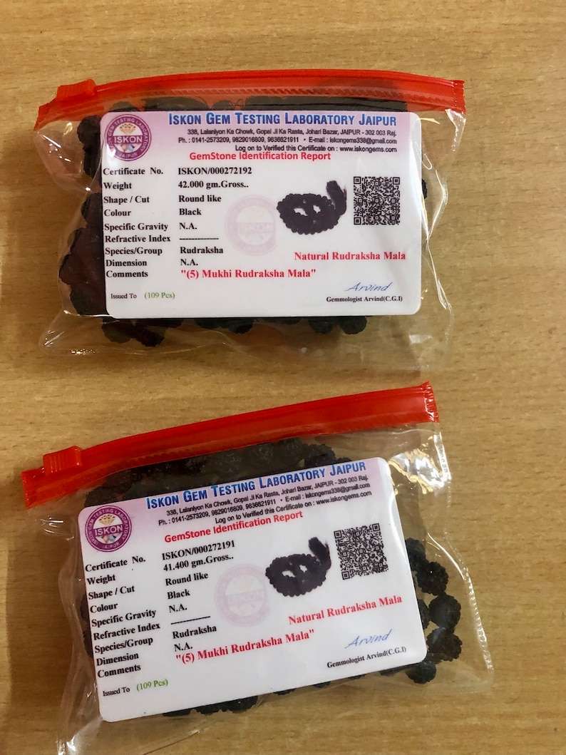 May include: A close-up of a small plastic bag containing a black beaded mala necklace. The bag has a white label with the text "ISKON GEM TESTING LABORATORY JAIPUR" and "GemStone Identification Report". The label also includes the certificate number "ISKON/000272192", weight "42.000 gm.Gross.", shape/cut "Round like", color "Black", specific gravity "N.A.", species/group "Rudraksha", dimension "N.A.", and comments "(5) Mukhi Rudraksha Mala".
