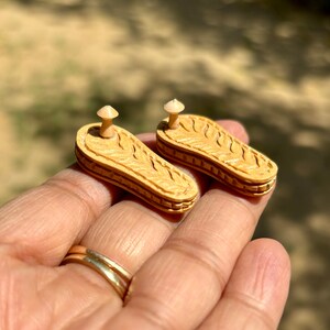 May include: Two small wooden sandals with a carved design and a small wooden knob on top. The sandals are light brown in color and are held in a hand.