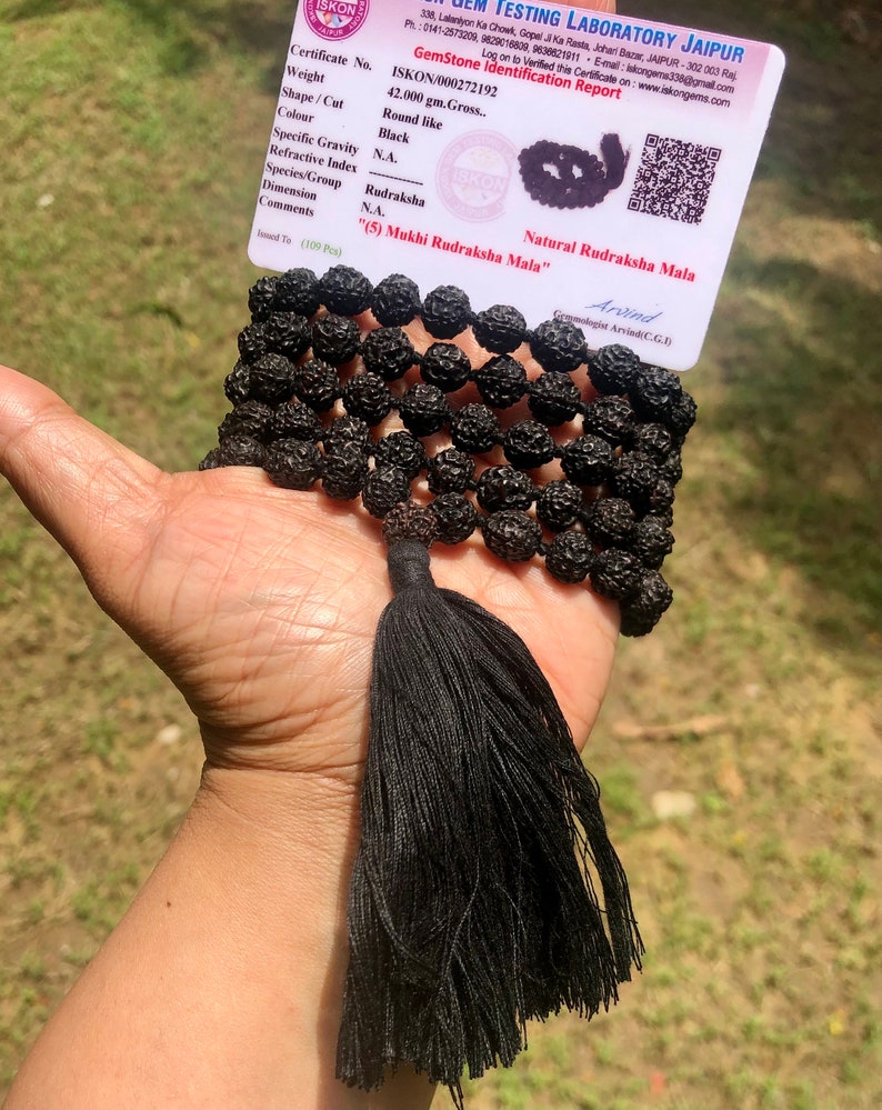 May include: A black Rudraksha Mala with a tassel, consisting of 109 beads. The mala is accompanied by a certificate of authenticity from ISKON Testing Laboratory Jaipur, stating the weight, shape, color, and other details of the mala. The certificate also includes the text "(5) Mukhi Rudraksha Mala" and "Natural Rudraksha Mala".
