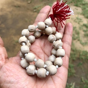 May include: A white wooden mala bead necklace with a red and white tassel. The necklace is made of round beads and is strung together with white thread.