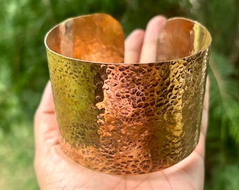 Könnte beinhalten: Ein breites, gehämmertes Kupferarmband. Das Armband hat eine strukturierte Oberfläche mit einer Mischung aus Gold- und Kupfertönen. Das offene Manschettendesign ermöglicht ein einfaches Tragen.