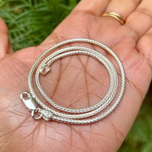 Puede incluir: Un collar de cadena de serpiente de plata con cierre de mosquetón. La cadena está hecha de pequeños eslabones entrelazados que crean un aspecto suave y fluido.