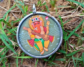 May include: A round silver pendant with a colorful image of the Hindu deity Ganesha playing a stringed instrument. The pendant is surrounded by a silver frame and has a loop at the top for attaching a chain.
