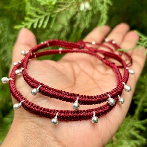 Pode incluir: Duas pulseiras de cordão trançado cor de vinho com contas de prata penduradas nas pontas. As pulseiras estão sobrepostas.