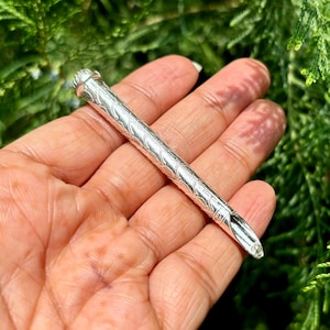 Puede incluir: Una herramienta de metal plateado y ornamentada, con una cabeza redondeada y una punta afilada. La herramienta presenta un patrón detallado y repetitivo a lo largo de su longitud. El objeto se sostiene en una mano sobre un fondo de follaje verde.