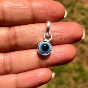 Puede incluir: Un amuleto de plata con un dise&ntilde;o de ojo azul malvado. El amuleto es redondo y tiene un borde blanco alrededor del ojo azul. La pupila del ojo es negra.