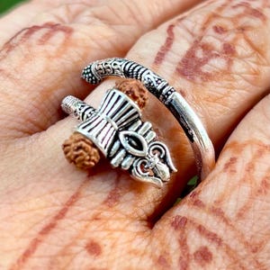 Peut inclure: Une bague argentée au design orné, avec un élément décoratif central et une graine naturelle. La bague s'enroule autour du doigt, mettant en valeur des détails et des textures complexes.