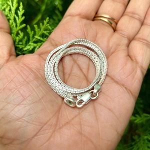 May include: A silver chain necklace with a box chain design. The chain is folded in half and held in a hand.