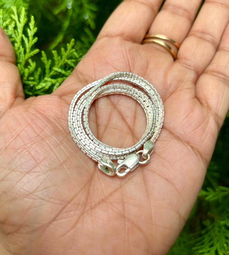 May include: A silver chain necklace with a box chain design. The chain is folded in half and held in a hand.