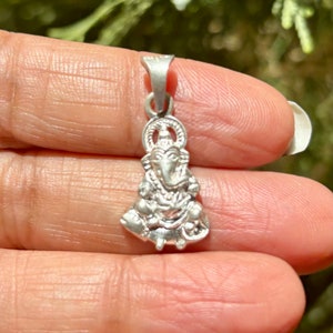 May include: Silver pendant featuring a detailed depiction of the Hindu deity Ganesha. The pendant is intricately crafted with a three-dimensional design and a loop at the top for attaching to a chain.