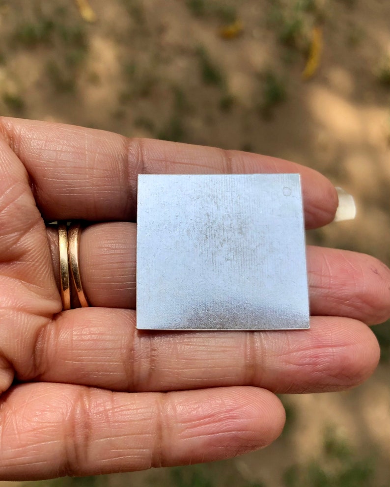 May include: A small, square, silver-colored metal plate with a slightly textured surface. The plate has a small, round hole in the upper right corner.