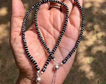 May include: Two black beaded anklets with silver charms. Each anklet has a small silver ball at the end of a chain.