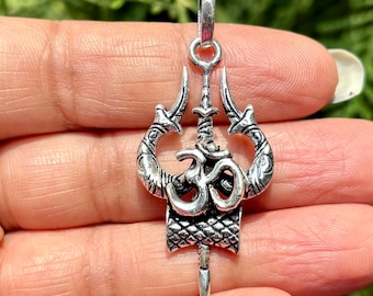 May include: Silver-toned pendant featuring a trident design with the Om symbol at the center. The pendant has a loop at the top for a chain and a decorative, textured pattern. The pendant is held in a hand against a blurred green background.