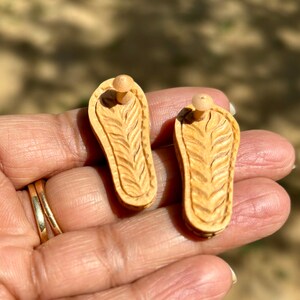 May include: A pair of wooden earrings shaped like sandals with a carved design. The earrings are a light brown color and have a small wooden post for attaching to the ear.