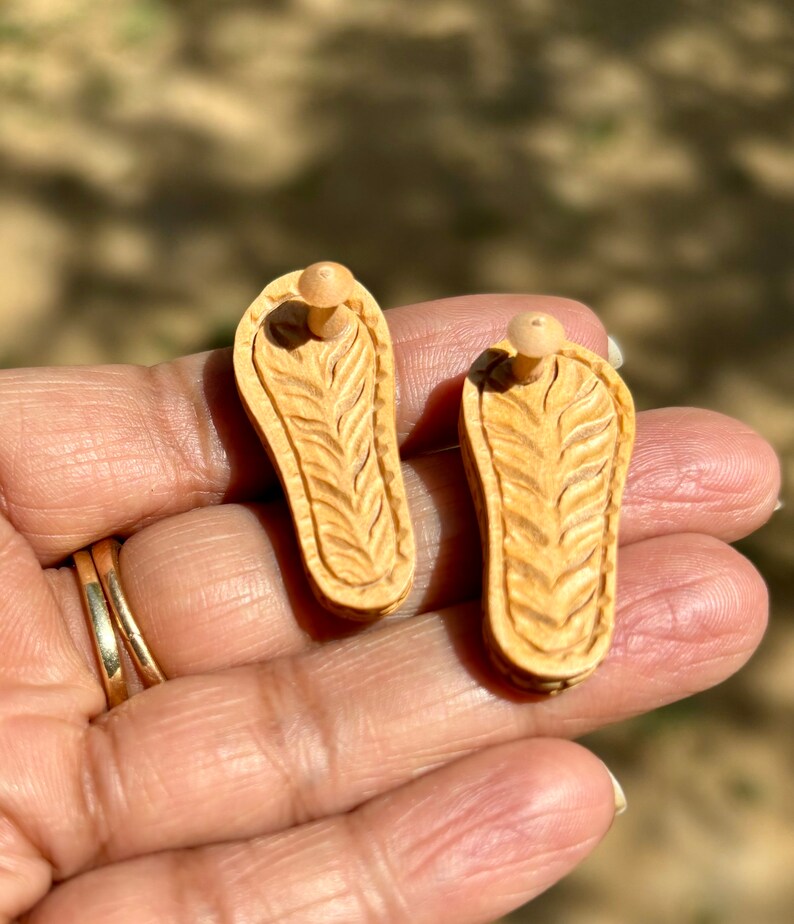 May include: A pair of wooden earrings shaped like sandals with a carved design. The earrings are a light brown color and have a small wooden post for attaching to the ear.