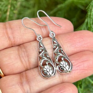 May include: A pair of silver dangle earrings with a delicate floral design. The earrings are teardrop shaped and have a filigree pattern.