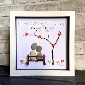 Könnte beinhalten: Ein weiß gerahmtes Bild mit weißem Hintergrund. Das Bild zeigt ein Paar aus grauen Steinen, das auf einer Bank aus Zweigen sitzt. Das Paar befindet sich unter einem Zweigbaum mit rosa Blüten. Der Text "Diamond Wedding Anniversary Phyllis & Bobby 01-03-1962." ist oben im Bild in Schwarz geschrieben.