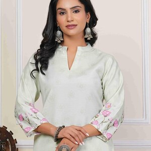 May include: A light green tunic with a floral pattern and a mandarin collar. The sleeves are adorned with pink floral designs. The model is wearing silver earrings, a bracelet, and a large ring. This outfit is suitable for casual or semi-formal occasions.